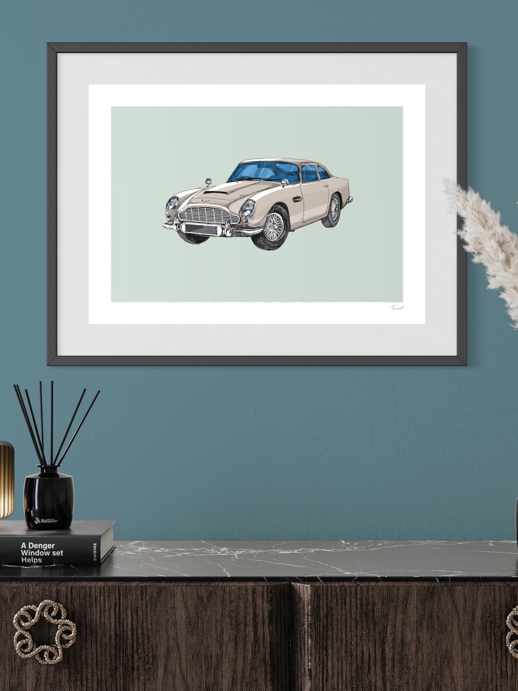 This image shows a giclee wall art print by Tom Laird Illustration of the British classic car, the Aston Martin DB5, framed in a beautiful living room with tasteful modern home decor. This art print of a silver example is available from Tom Laird Illustration in a variety of sizes.